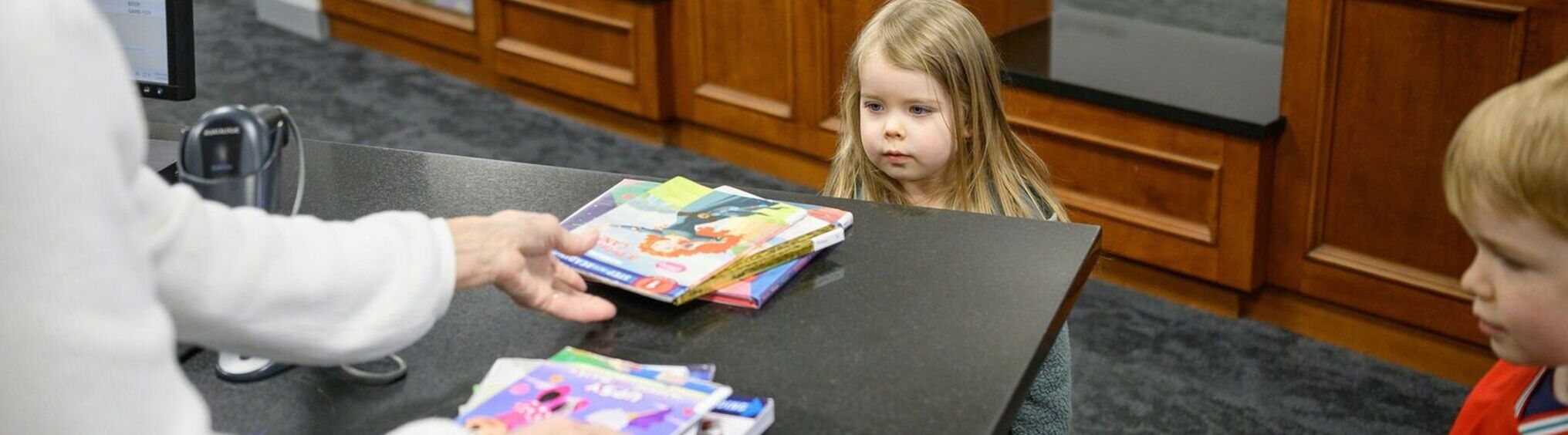 Two children waiting at checkout counter while library employee checks out books.