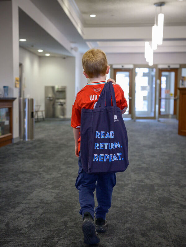 Child walking towards library exit holding library bag that reads, 