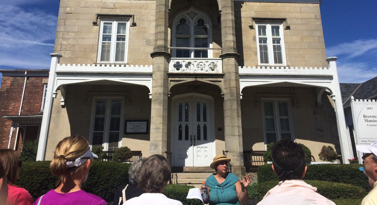 Staff member standing and speaking in front of a historic home in Hudson while a crowd of people listen during a walking tour.